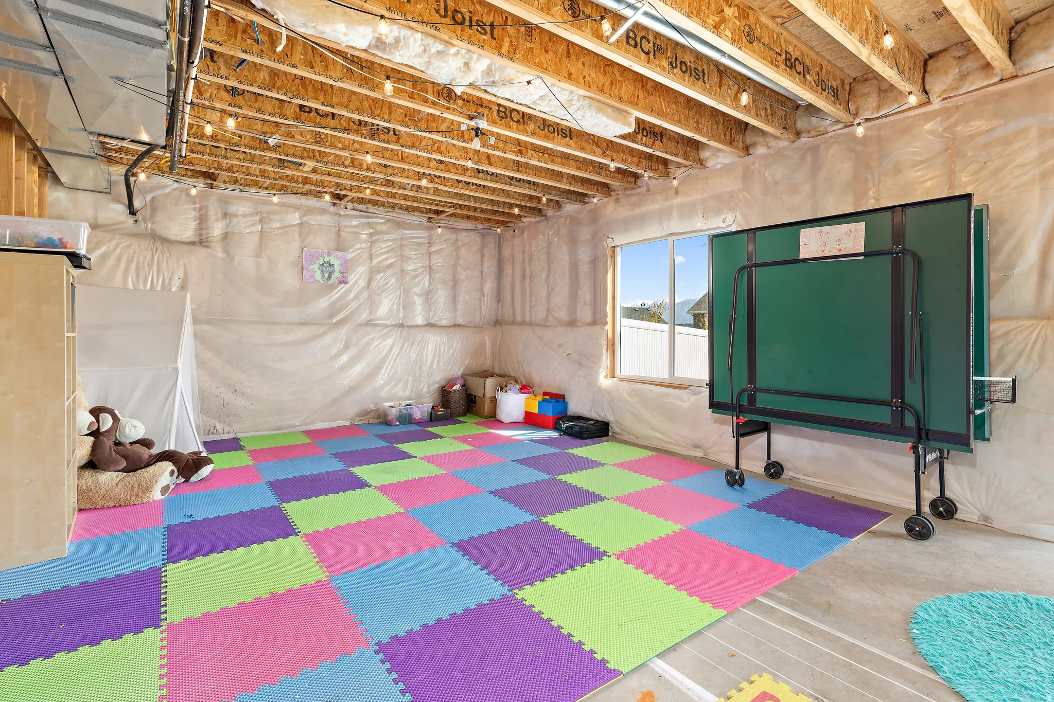 Unfinished basement space featuring exposed wood joists, a large window, and string lighting