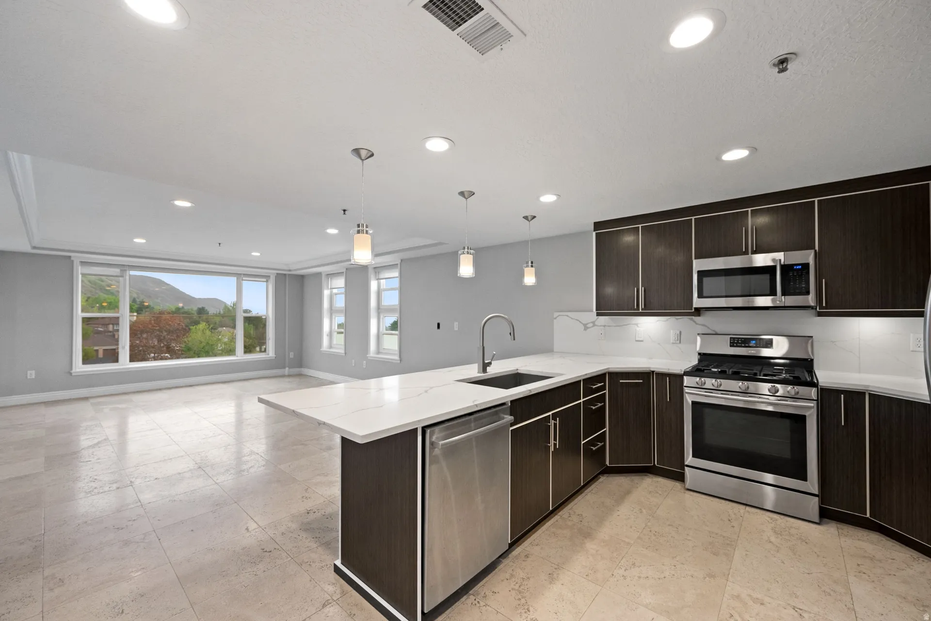 Kitchen with dark wood finish cabinetry, stainless steel appliances, light stone counters, a peninsula, and hanging light fixtures