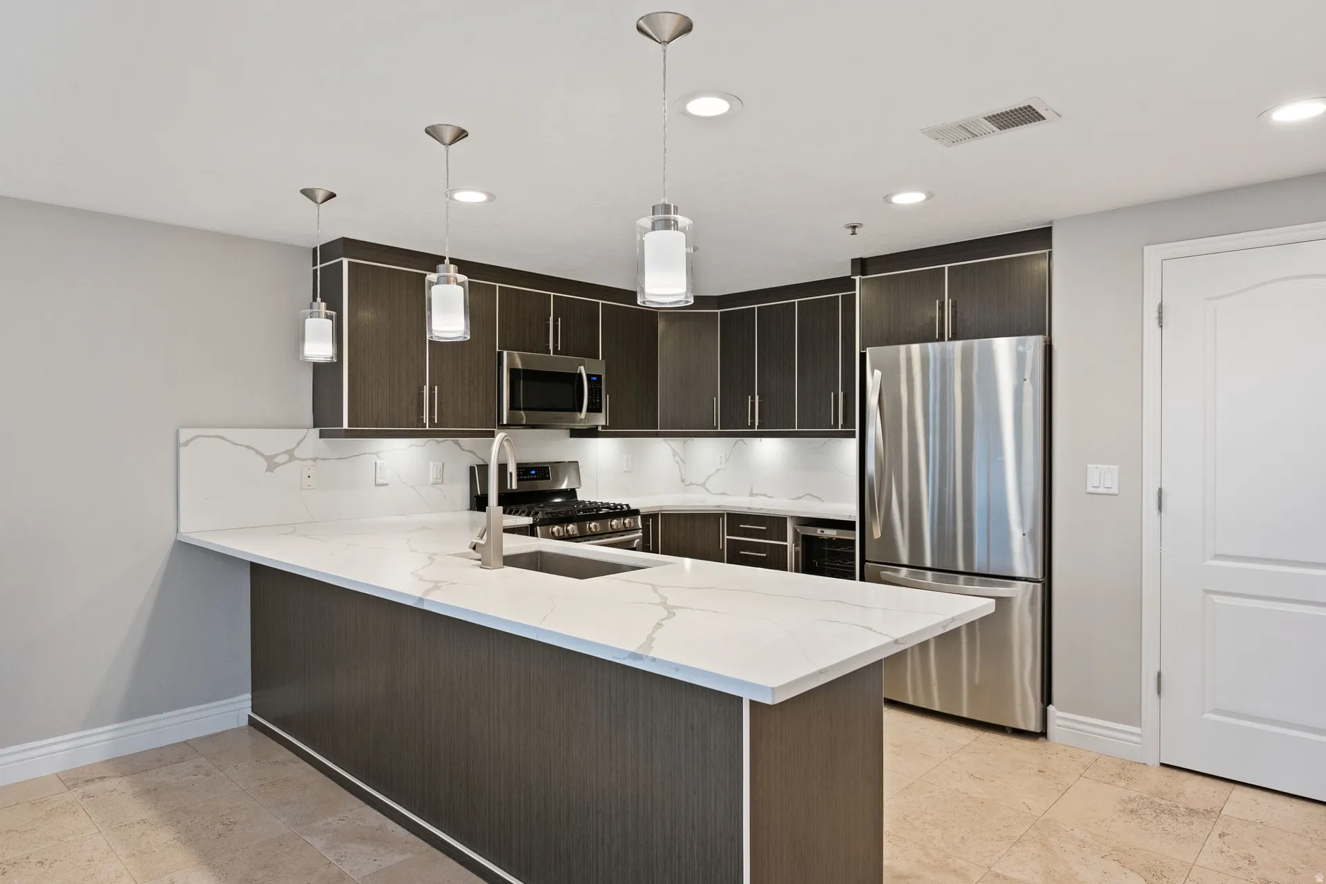 Kitchen featuring dark wood finish cabinets, stainless steel appliances, light stone countertops, a peninsula, and hanging light fixtures