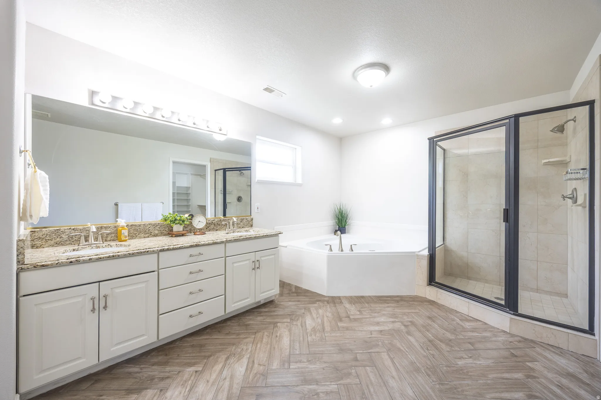 Spacious bathroom featuring a dual vanity with a granite countertop, a large walk-in shower with a glass enclosure, a corner soaking tub, and wood-finish flooring