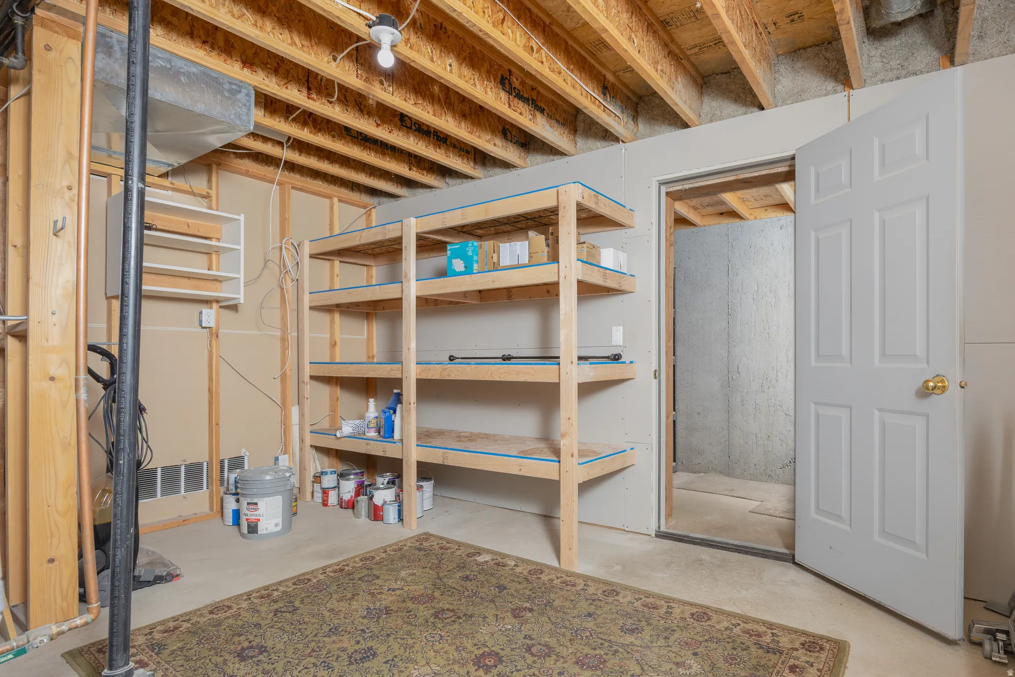 Unfinished basement space featuring exposed wood joists and subflooring, built-in wooden shelving, and a panel door leading to an additional unfinished area