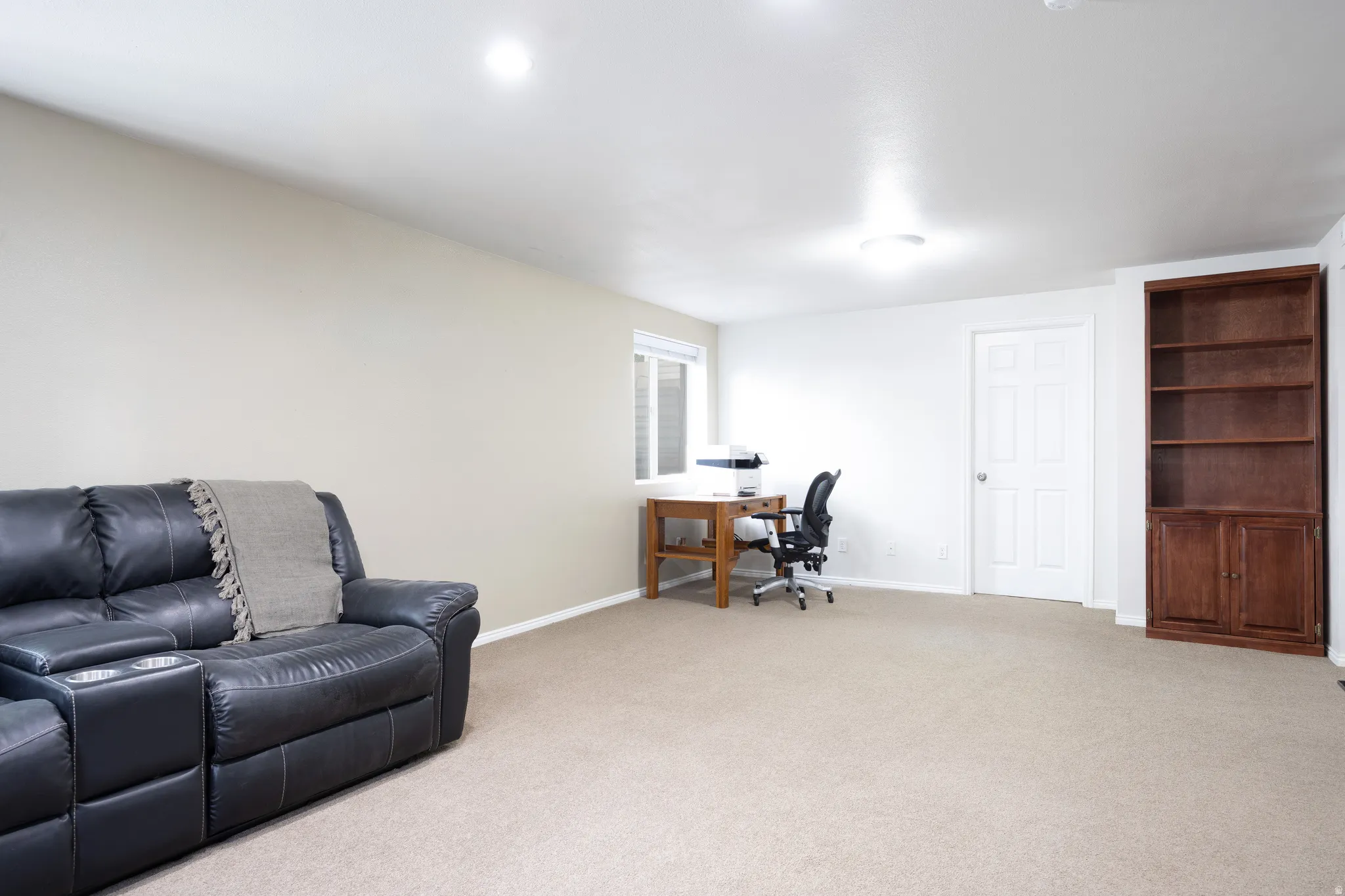 Spacious multi-purpose room featuring plush carpet flooring, recessed ceiling lighting, and a built-in wood-finish shelving unit