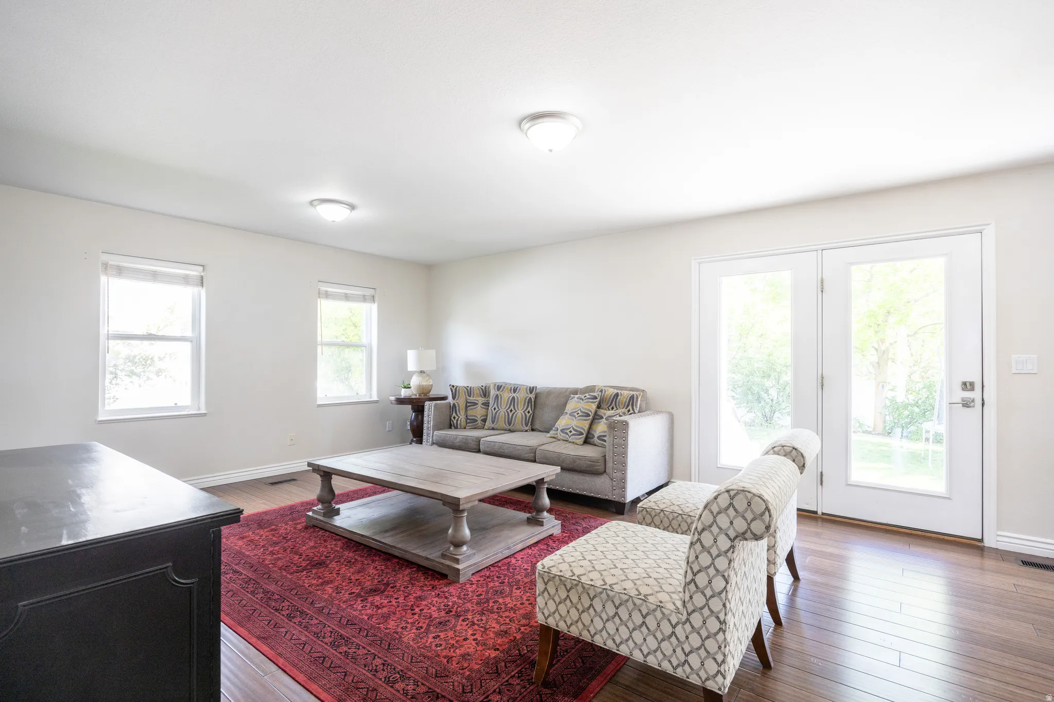 Living area featuring wood-finish flooring, light neutral wall tones, two windows with blinds, two ceiling-mounted light fixtures, and white French doors