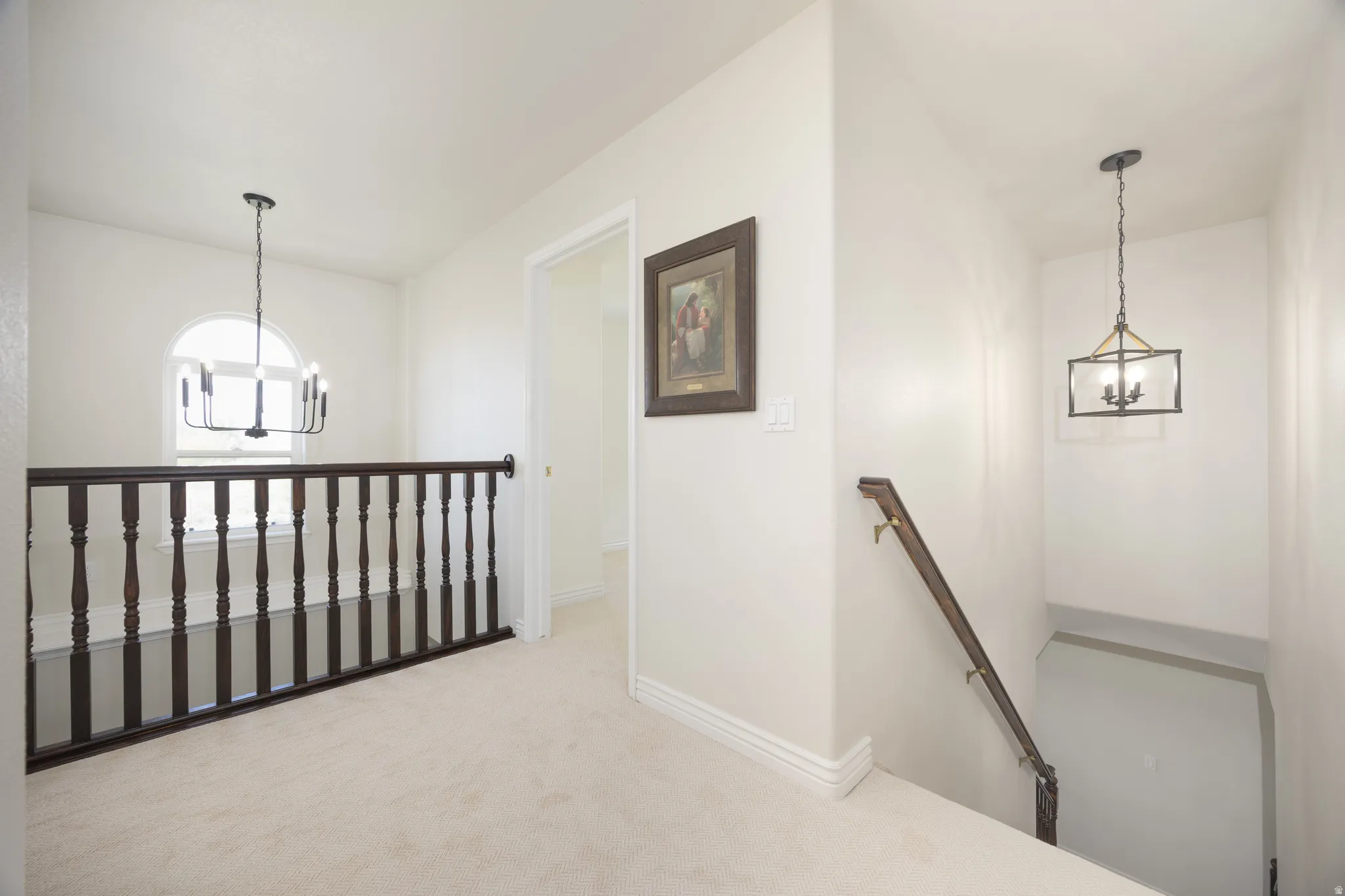 Upper-level landing featuring light-toned carpeting, a dark wood baluster railing, an arched window, and two distinct overhead light fixtures