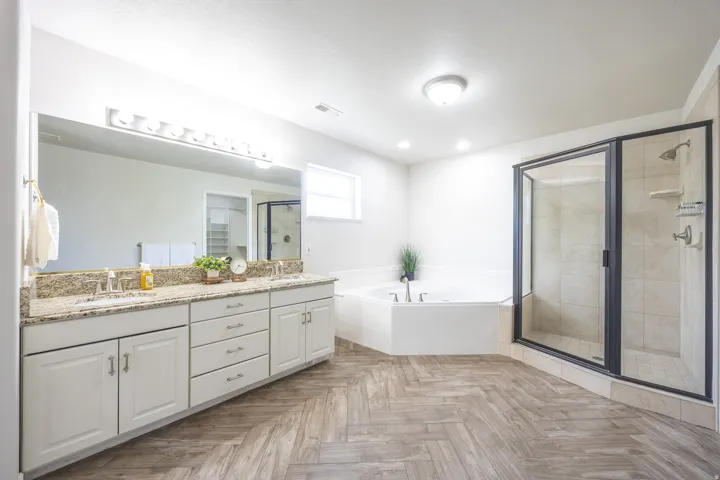 Spacious bathroom featuring a dual vanity with a granite countertop, a large walk-in shower with a glass enclosure, a corner soaking tub, and wood-finish flooring