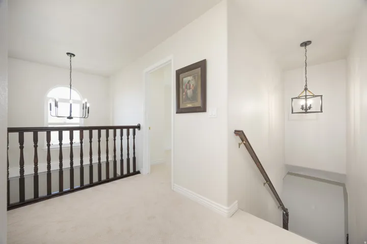 Upper-level landing featuring light-toned carpeting, a dark wood baluster railing, an arched window, and two distinct overhead light fixtures