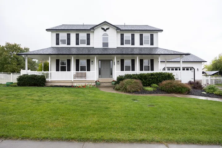 Colonial-style residence featuring a prominent covered front porch, white siding, black window shutters, a two-car garage, and established landscaping