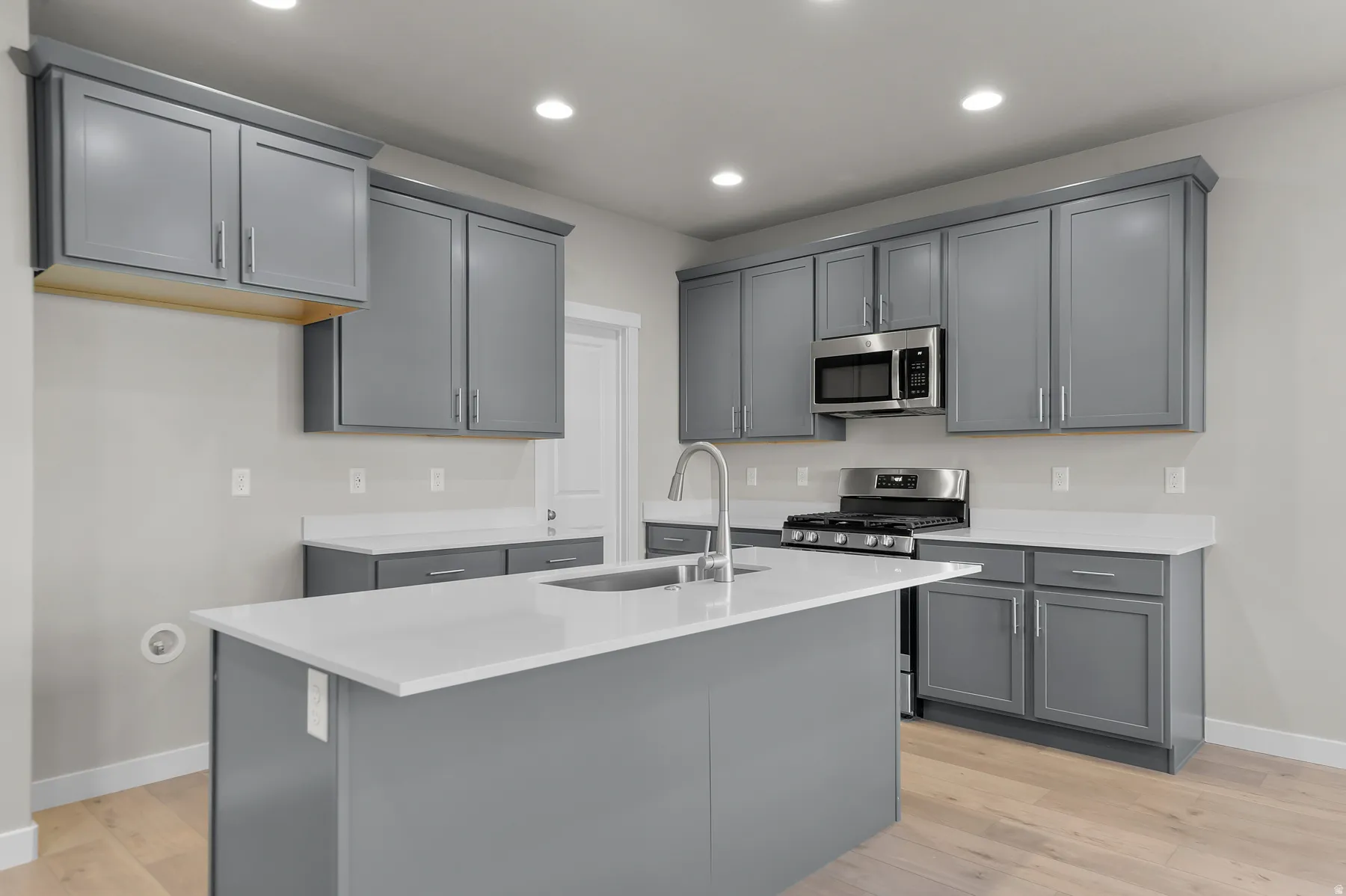 Modern kitchen featuring an island with an undermount sink, gray cabinetry, light-toned countertops, stainless steel appliances, and wood-finish flooring