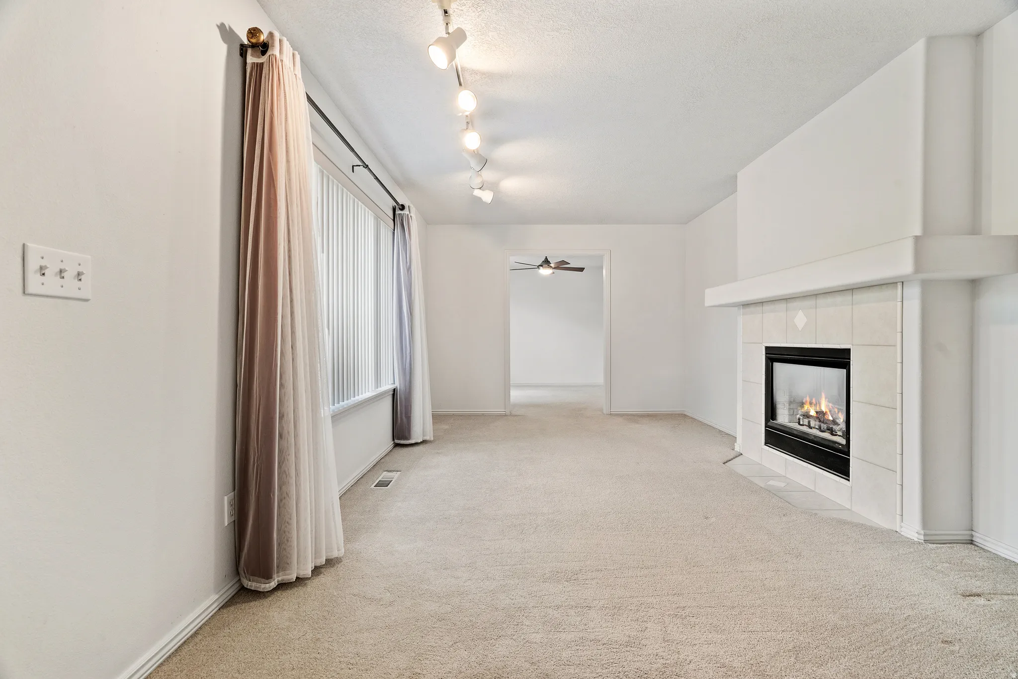 Spacious room featuring a fireplace with a tiled surround and mantel, track lighting, a large window with blinds, and neutral carpeting