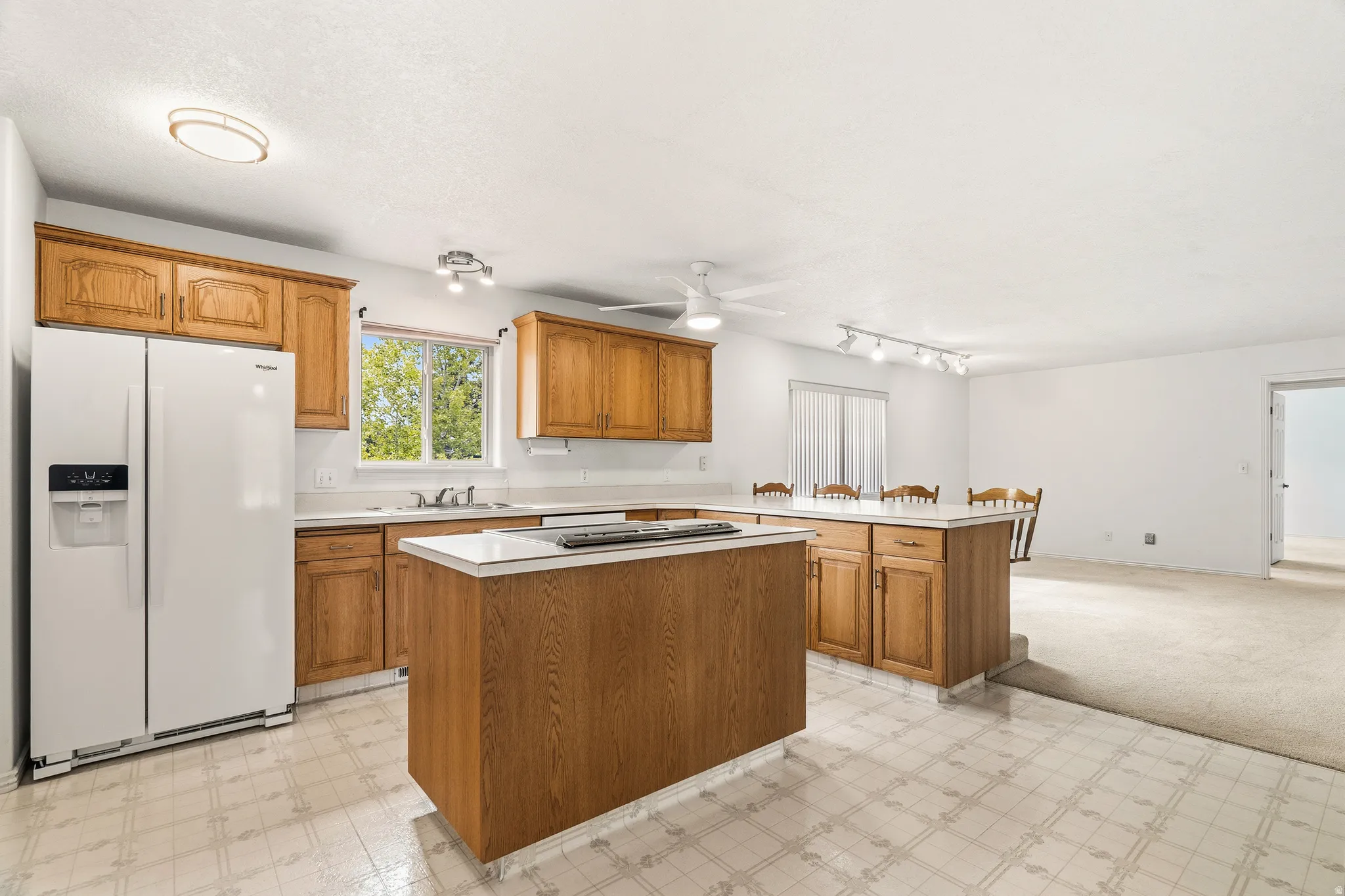 Spacious kitchen featuring a central island with a solid surface countertop, light wood-finish cabinetry, a white side-by-side refrigerator, a window over the sink, and tile-look flooring