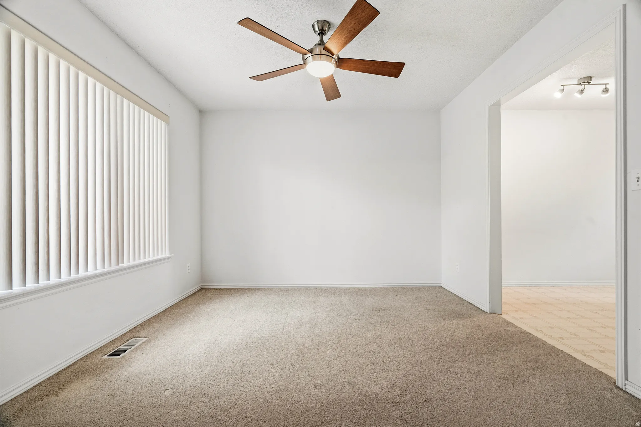 Carpeted room featuring a ceiling fan with wood-finish blades, a large window with vertical blinds, and white walls