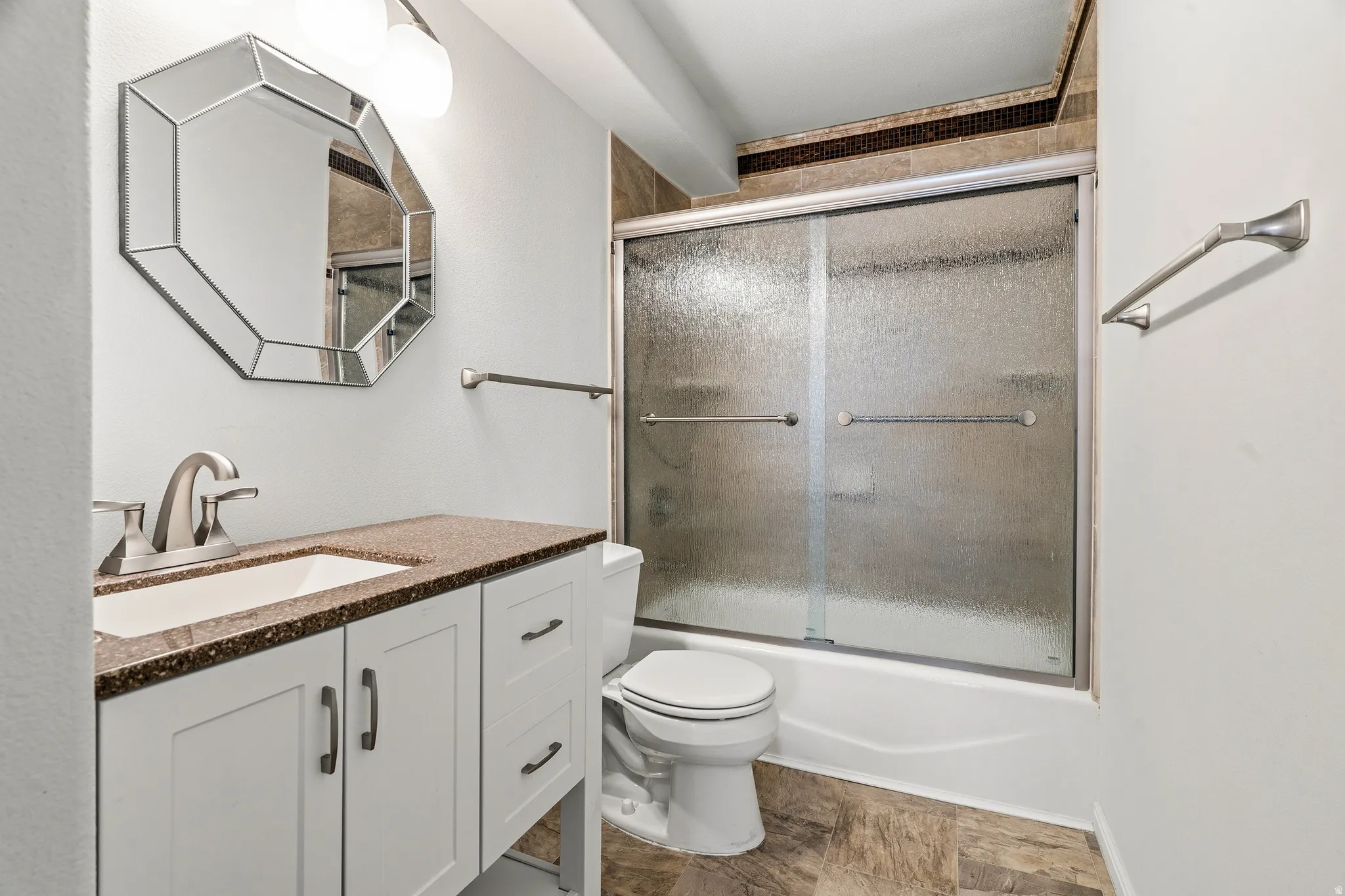 Bathroom featuring a vanity with a white basin and a speckled countertop, an octagonal mirror, a shower-over-tub with frosted sliding doors, and a tiled accent border