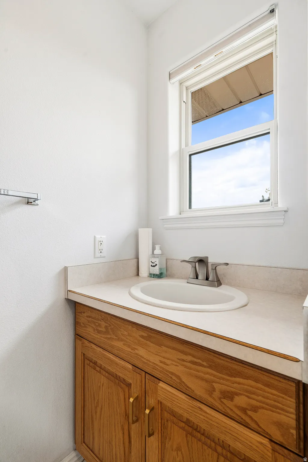 White single-hung window with a roller shade, wood-finish vanity with panel doors and brass-tone hardware, integrated sink, and brushed nickel faucet