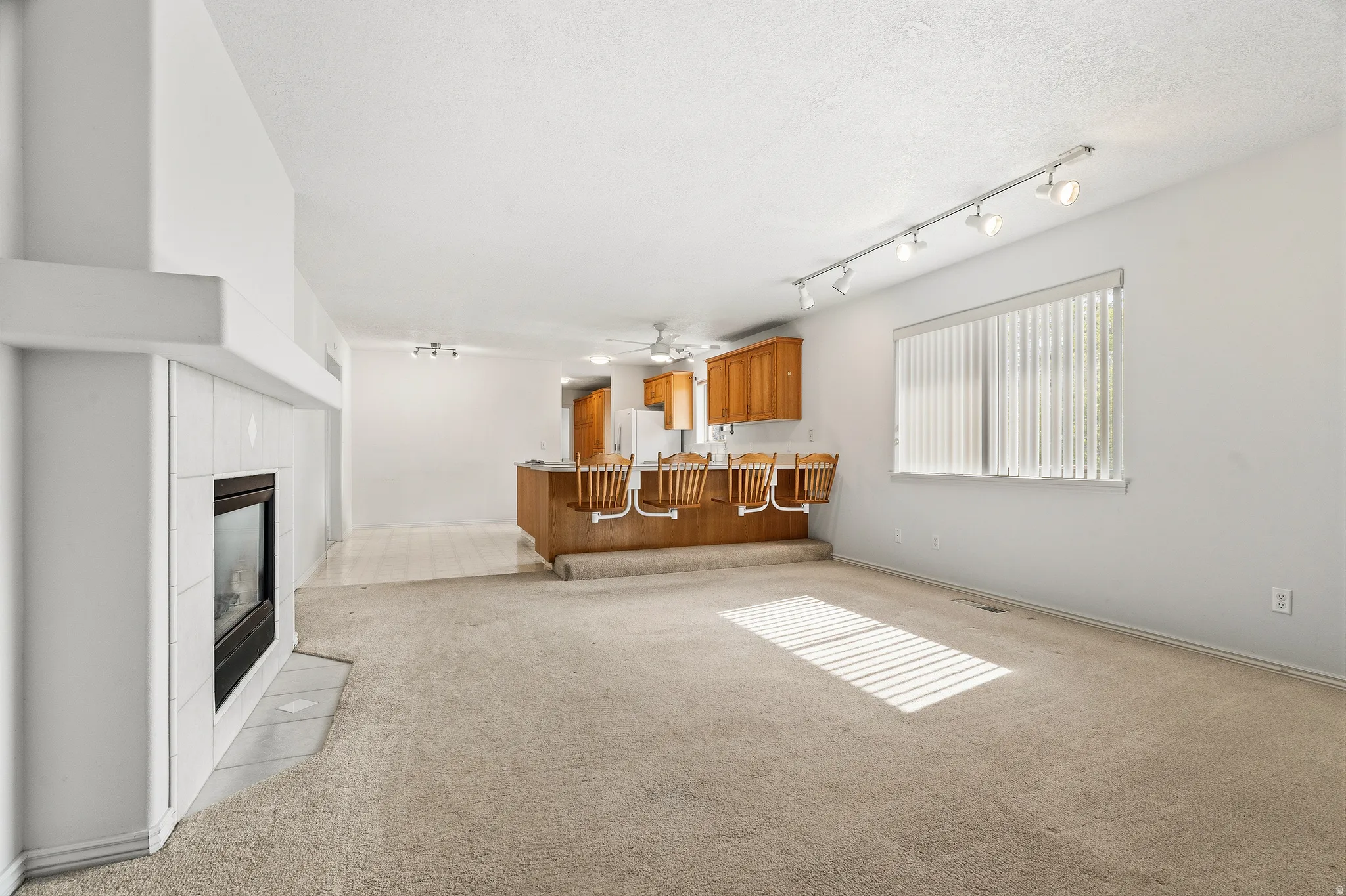 Spacious living area featuring a central fireplace with a tile surround, light-toned carpeting, and a large window with horizontal blinds