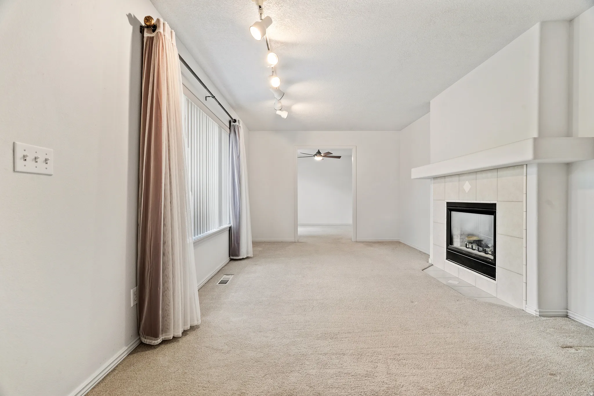Living area featuring a fireplace with a tiled surround and mantel, track lighting, and a large window with vertical blinds