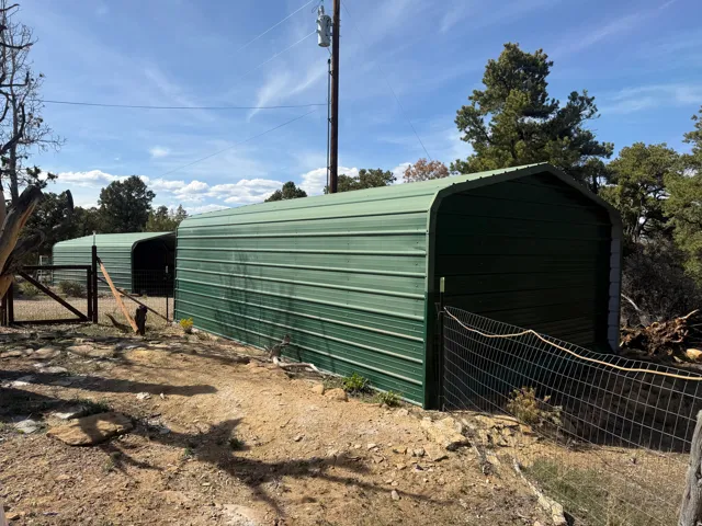Two green metal carports with a third partially visible, set on a natural terrain, featuring a wire fence and a wooden gate