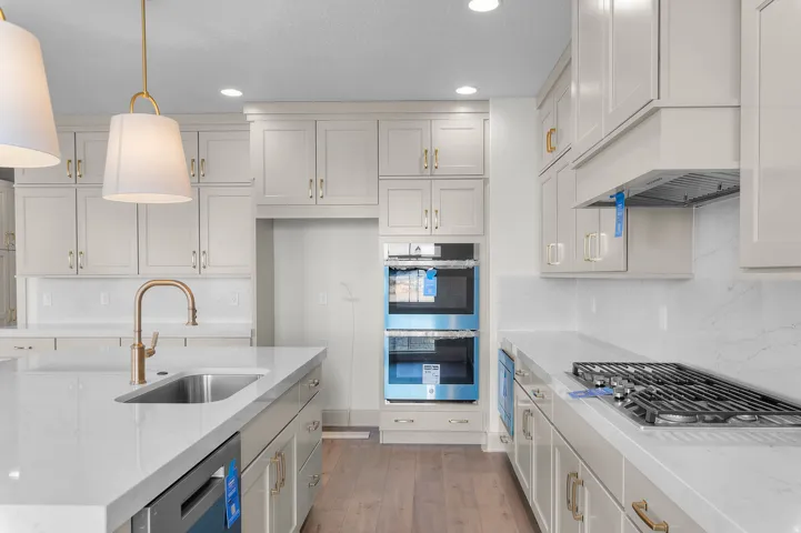 Contemporary kitchen featuring a large island with an undermount sink and polished brass faucet, white shaker cabinetry, integrated double wall ovens, a gas cooktop, and light wood-finish flooring