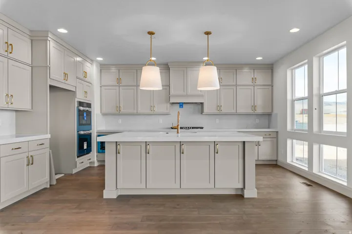 Spacious kitchen featuring an expansive center island, light-toned cabinetry with gold-finish hardware, built-in double ovens, wood-finish flooring, and multiple large windows