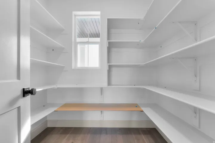 Spacious walk-in pantry featuring extensive white shelving, a natural wood-finish countertop, a single window, and dark wood-finish flooring