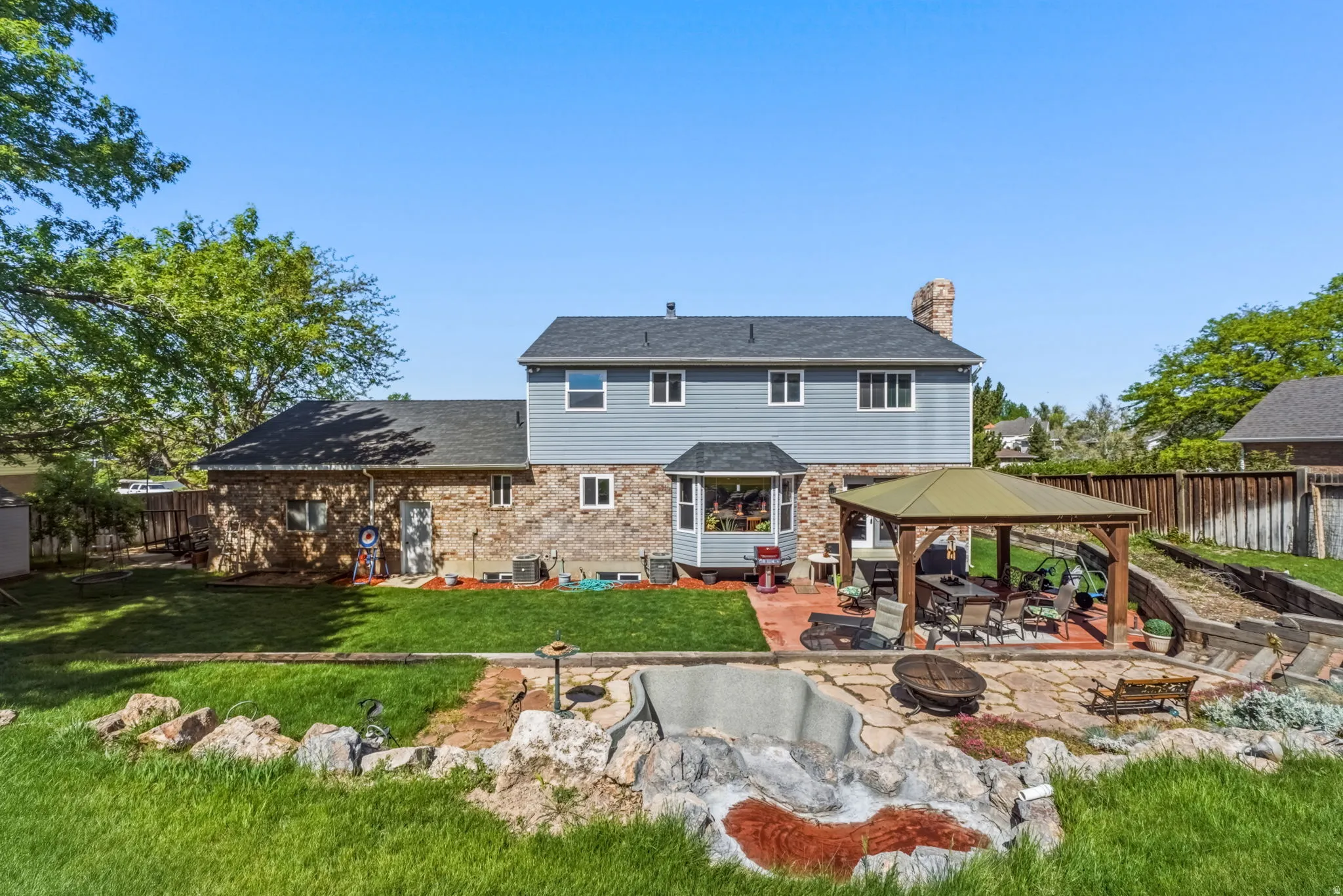 Rear exterior featuring stone and siding finishes, a multi-level patio with a built-in gazebo, a bay window, and a stone-lined water feature