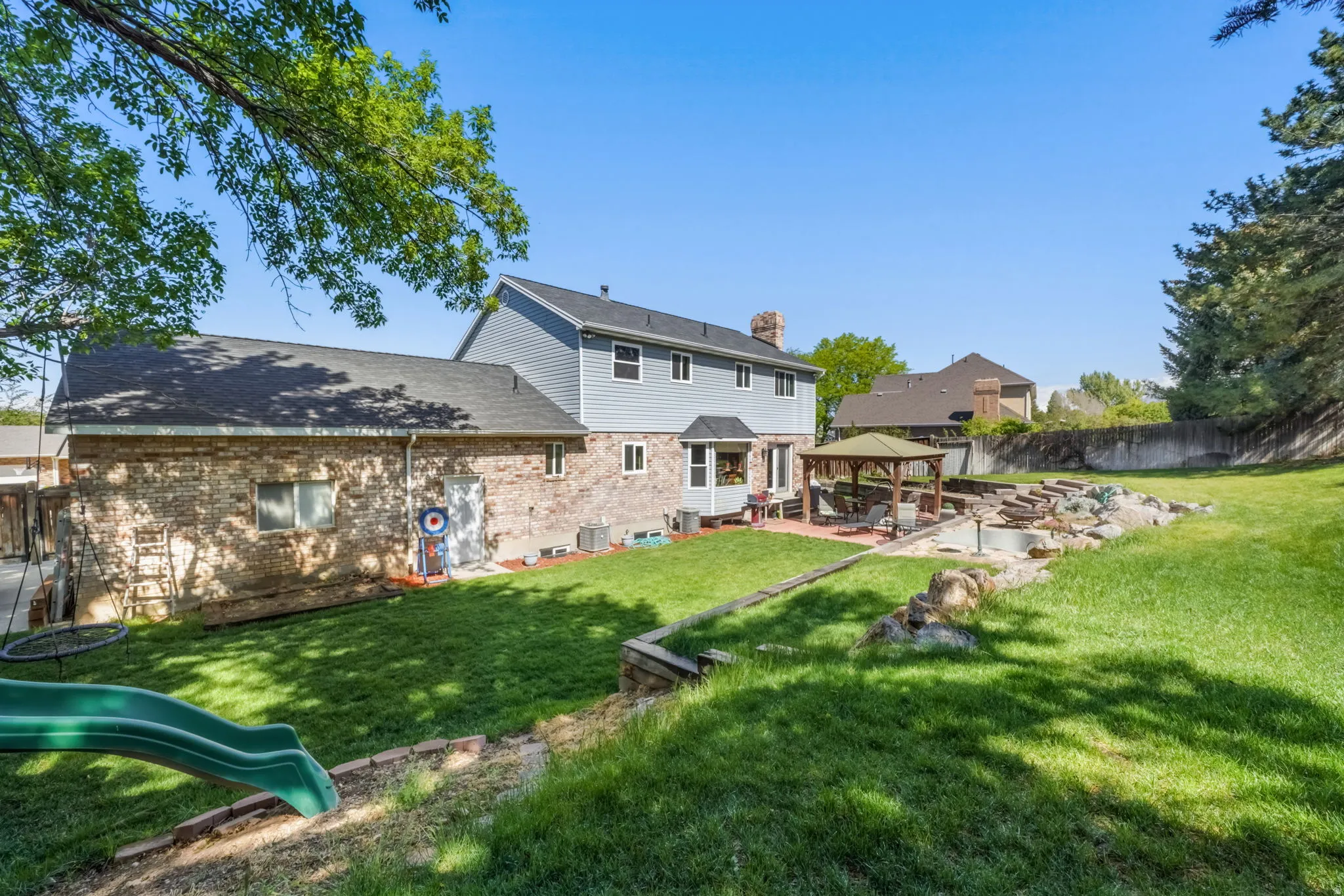 Expansive backyard featuring a manicured lawn, a multi-level wood-retaining wall, a stone patio, and a wood-frame gazebo