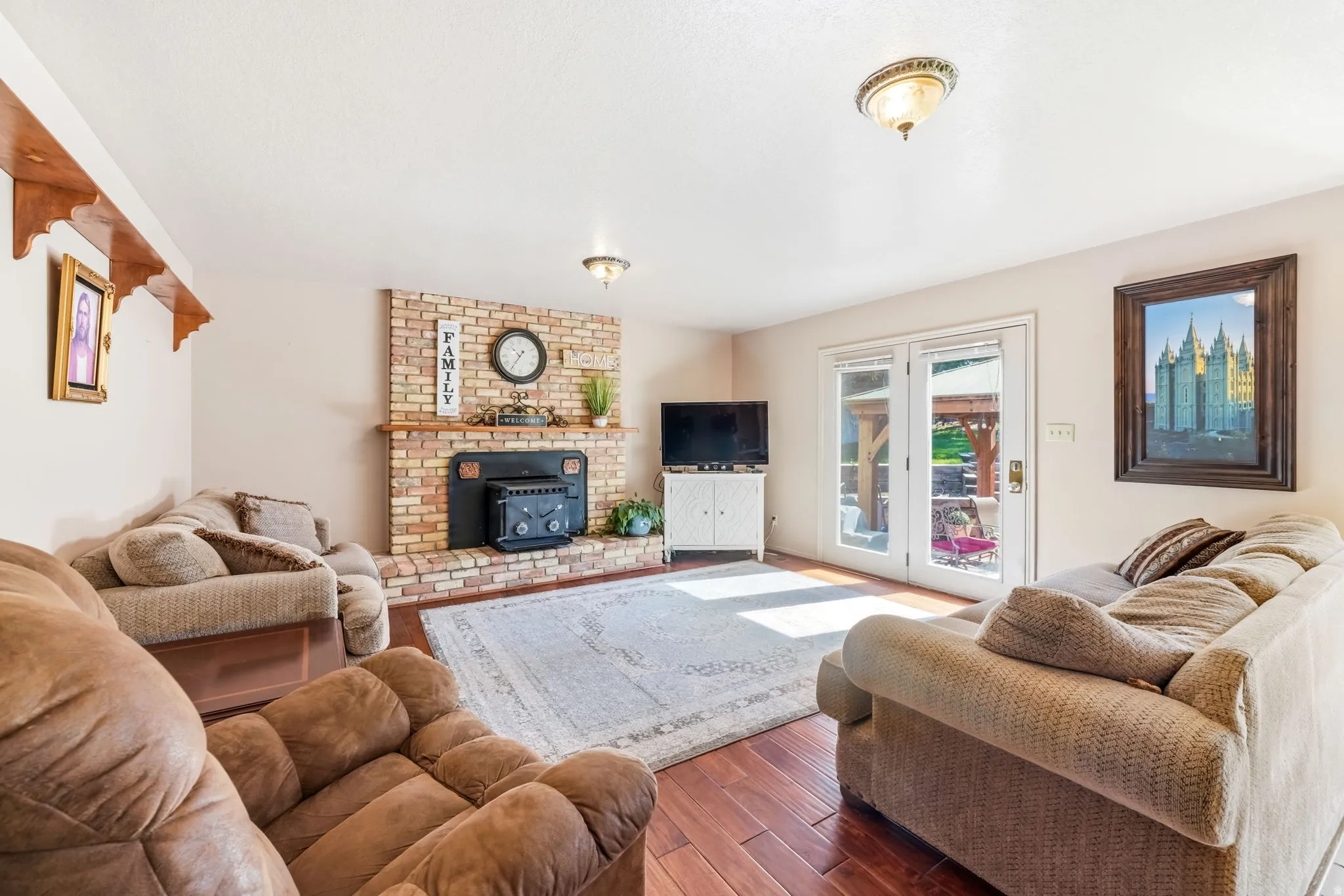 Living area featuring brick fireplace with wood mantel, a black wood-burning stove insert, wood-finish flooring, and white French doors leading to an outdoor space