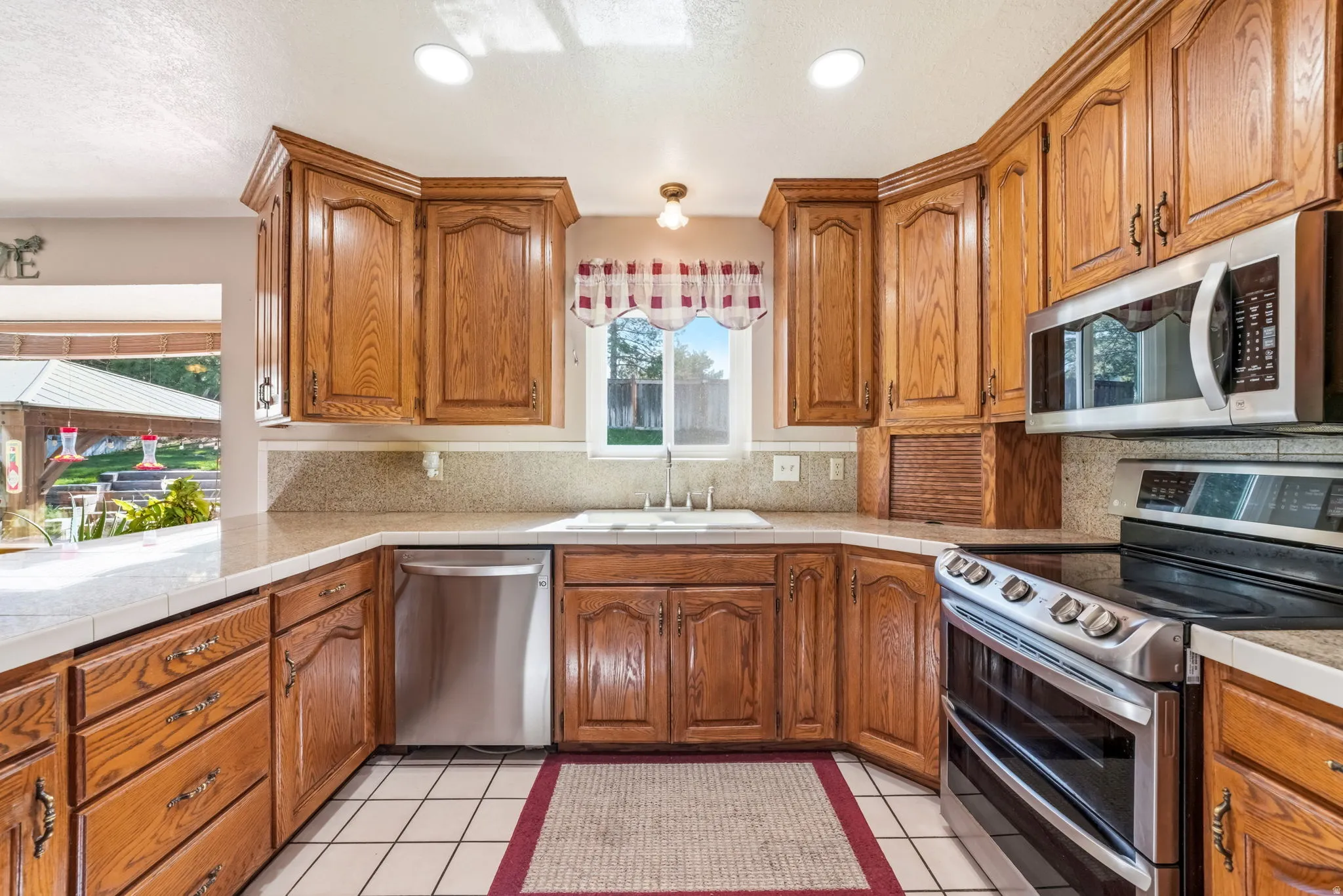 Kitchen featuring wood cabinetry, white tile flooring, stainless steel appliances, a white sink, and a window with exterior views