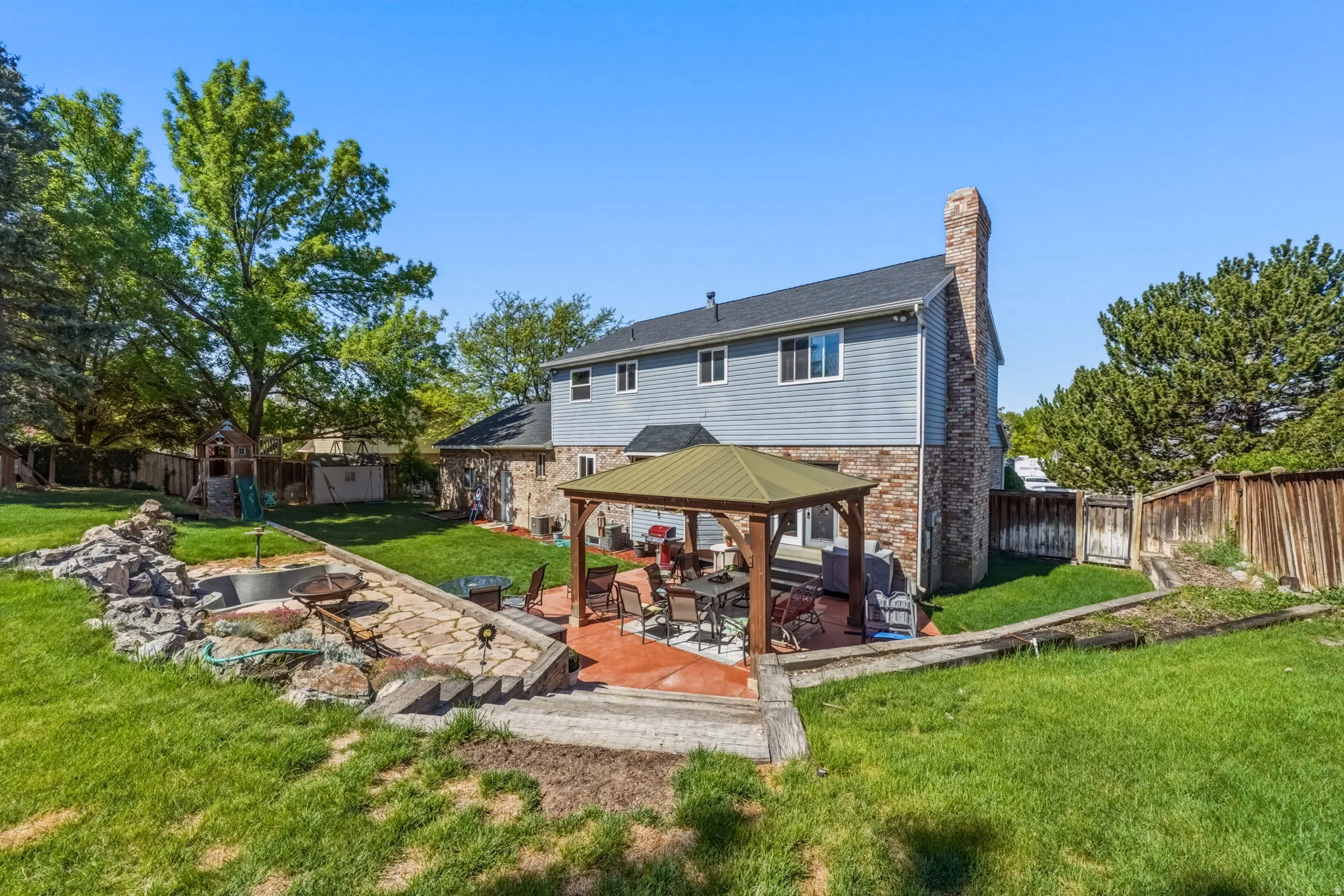 Spacious backyard featuring a stone patio with an outdoor gazebo, mature trees, and a terraced stone garden area