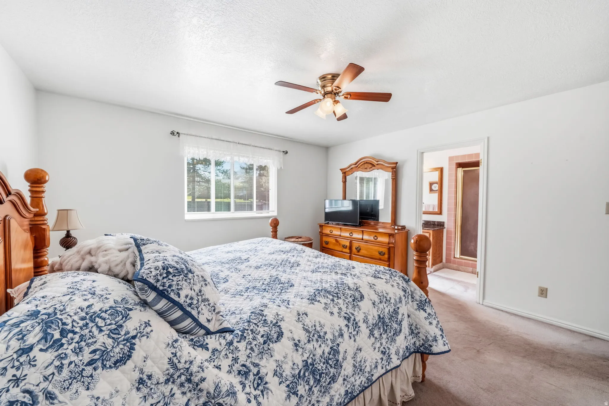 Carpeted bedroom featuring a ceiling fan with integrated lighting, a window with exterior views, and an open doorway leading to a bathroom with a shower enclosure