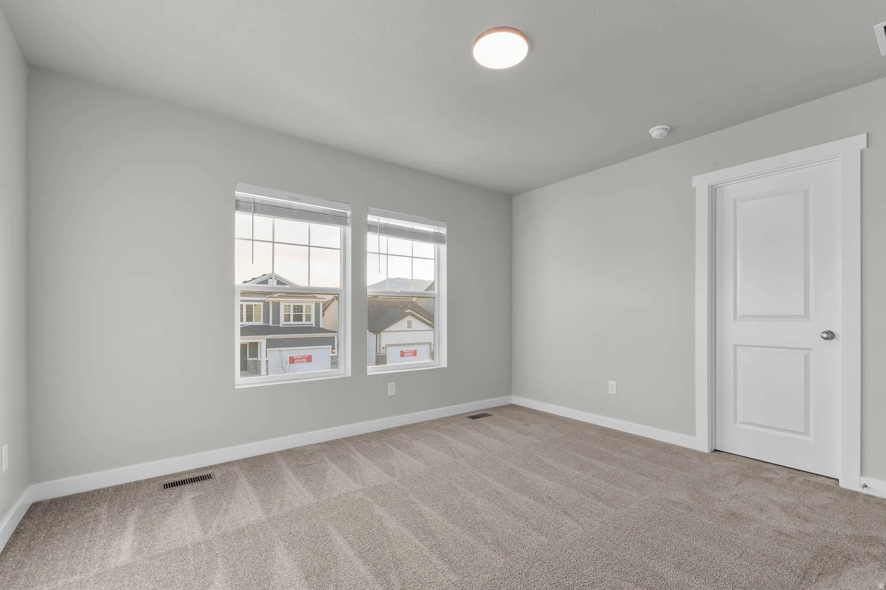 Light gray interior walls with white trim, two clear glass windows, neutral patterned carpet, white two-panel interior door, and a flush-mount ceiling light fixture