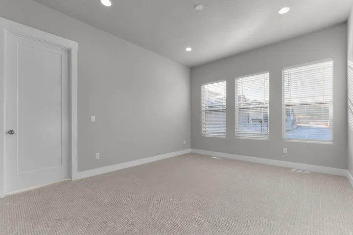 Spacious room with light gray walls, plush carpeting, three large windows with blinds, recessed lighting, and a paneled door