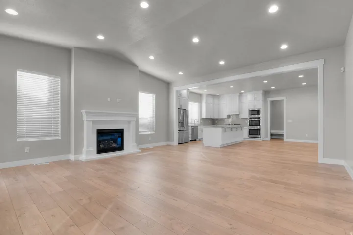 Spacious open-concept living area featuring light wood-finish flooring, a white mantel fireplace, vaulted ceilings with recessed lighting, and a kitchen with an island and stainless steel appliances
