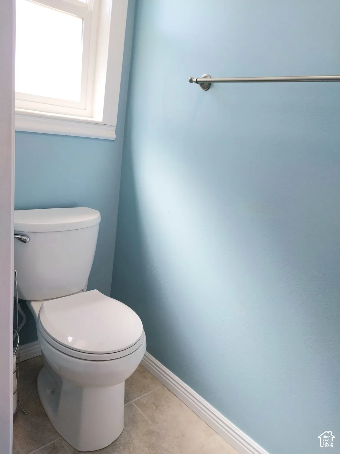 White toilet, window with white trim, light blue wall, tiled flooring, and a brushed nickel towel bar
