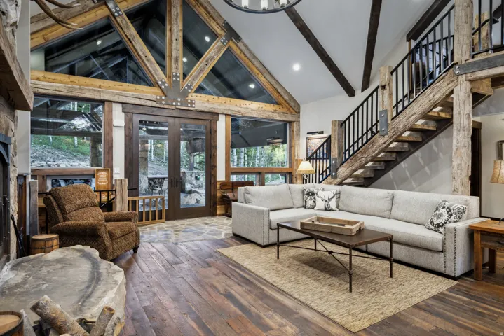 Vaulted ceiling with exposed wood beams and architectural glass, rich wood-finish flooring, stone fireplace, and a striking open staircase with iron railings