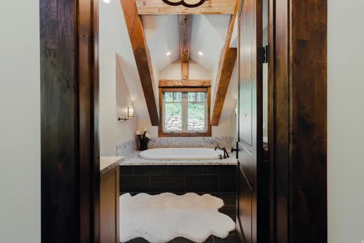 Primary Bathroom featuring vaulted ceilings with exposed timber beams, a natural light-filled window, dual vanity with stone countertop, and polished nickel fixtures beneath a statement pendant light fixture