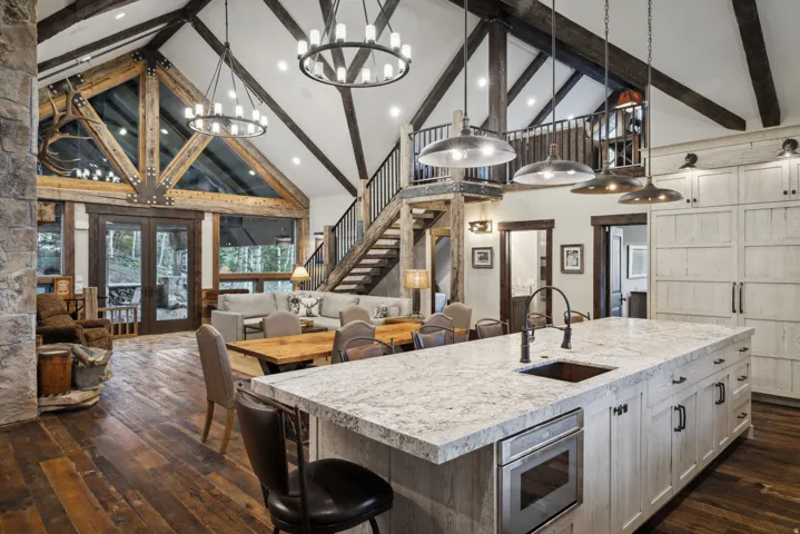 Vaulted ceiling with exposed wood beams, expansive floor-to-ceiling windows, wood-finish flooring, central kitchen island with integrated sink, and a rustic stone fireplace