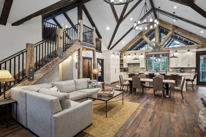 Vaulted ceiling with exposed wood beams, wood-finish flooring, custom wood and metal staircase, kitchen with upper and lower cabinetry, and expansive window with exterior scenery