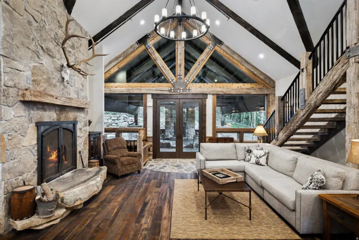 Vaulted ceiling with exposed wood beams, stone fireplace with a substantial hearth, wood-finish flooring, and a round chandelier