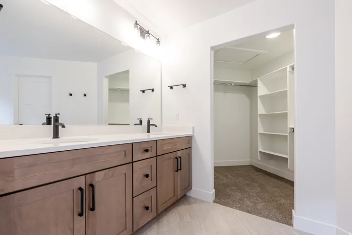 Dual vanity with matte black fixtures, light wood-finish cabinetry, and a spacious walk-in closet with built-in shelving
