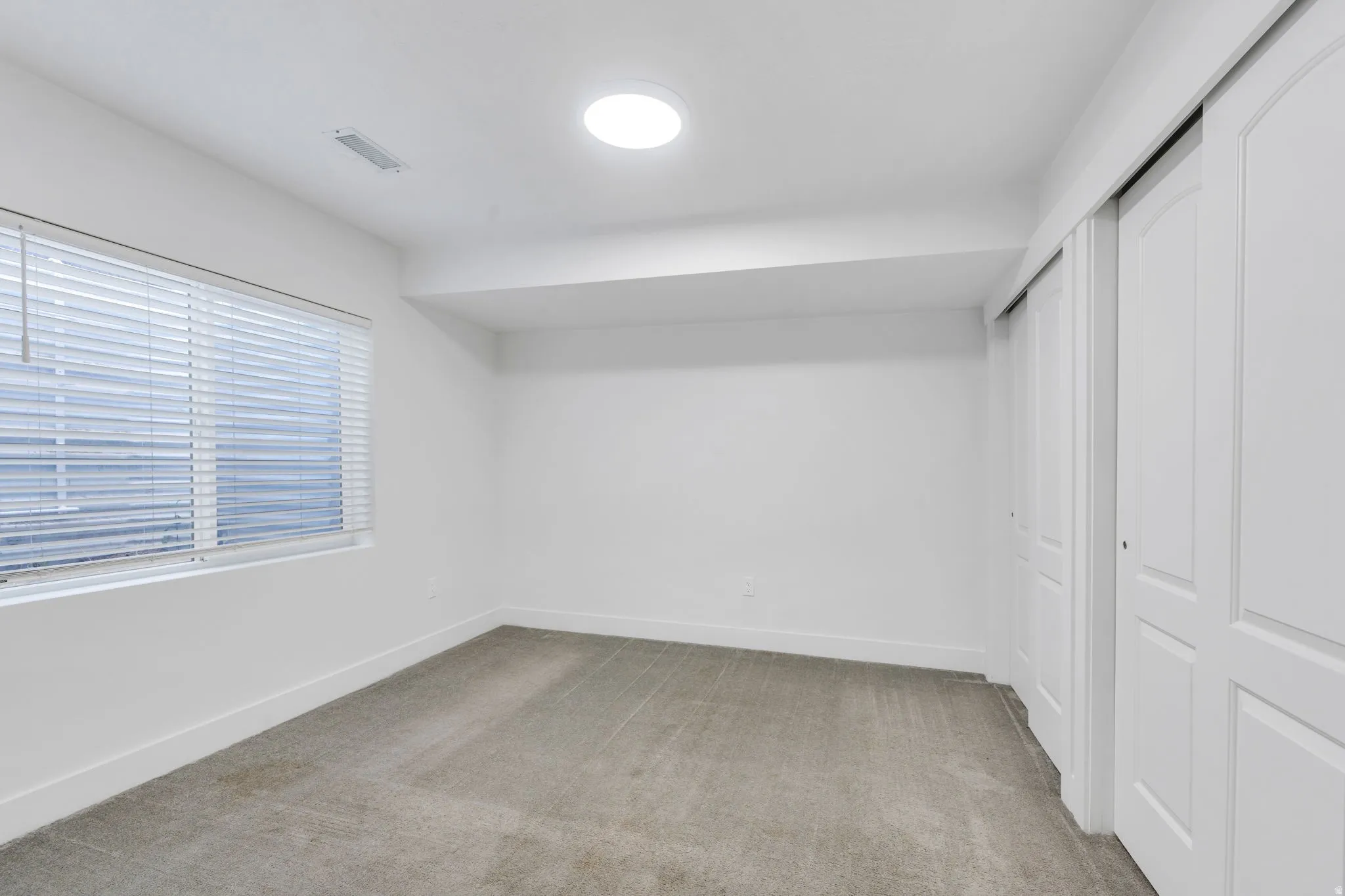 Bedroom featuring a large window with horizontal blinds, neutral-toned carpet, and a built-in closet with white bifold doors