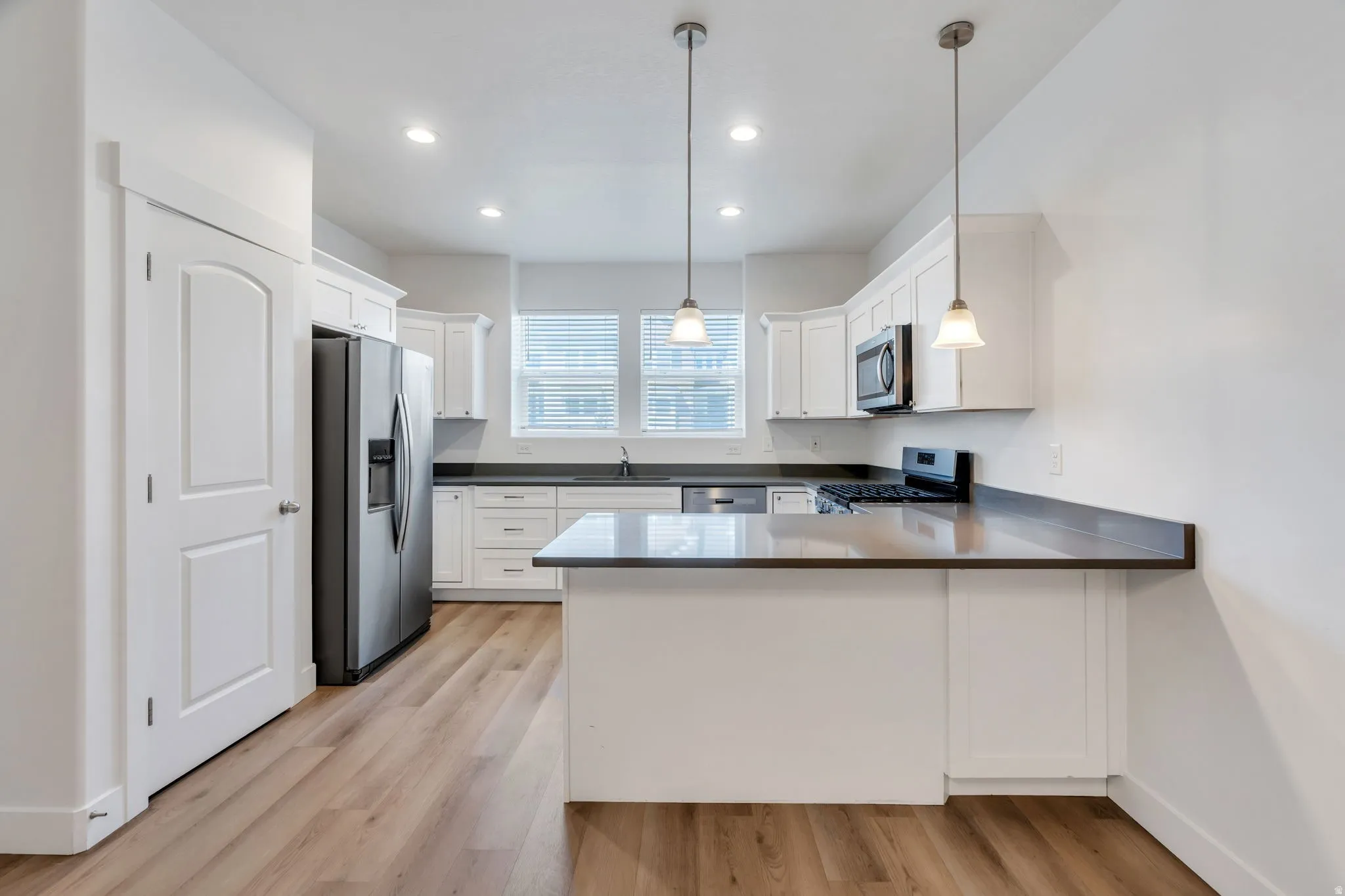 Modern kitchen featuring wood-finish flooring, white cabinetry, dark countertops, a stainless steel refrigerator, and pendant lighting