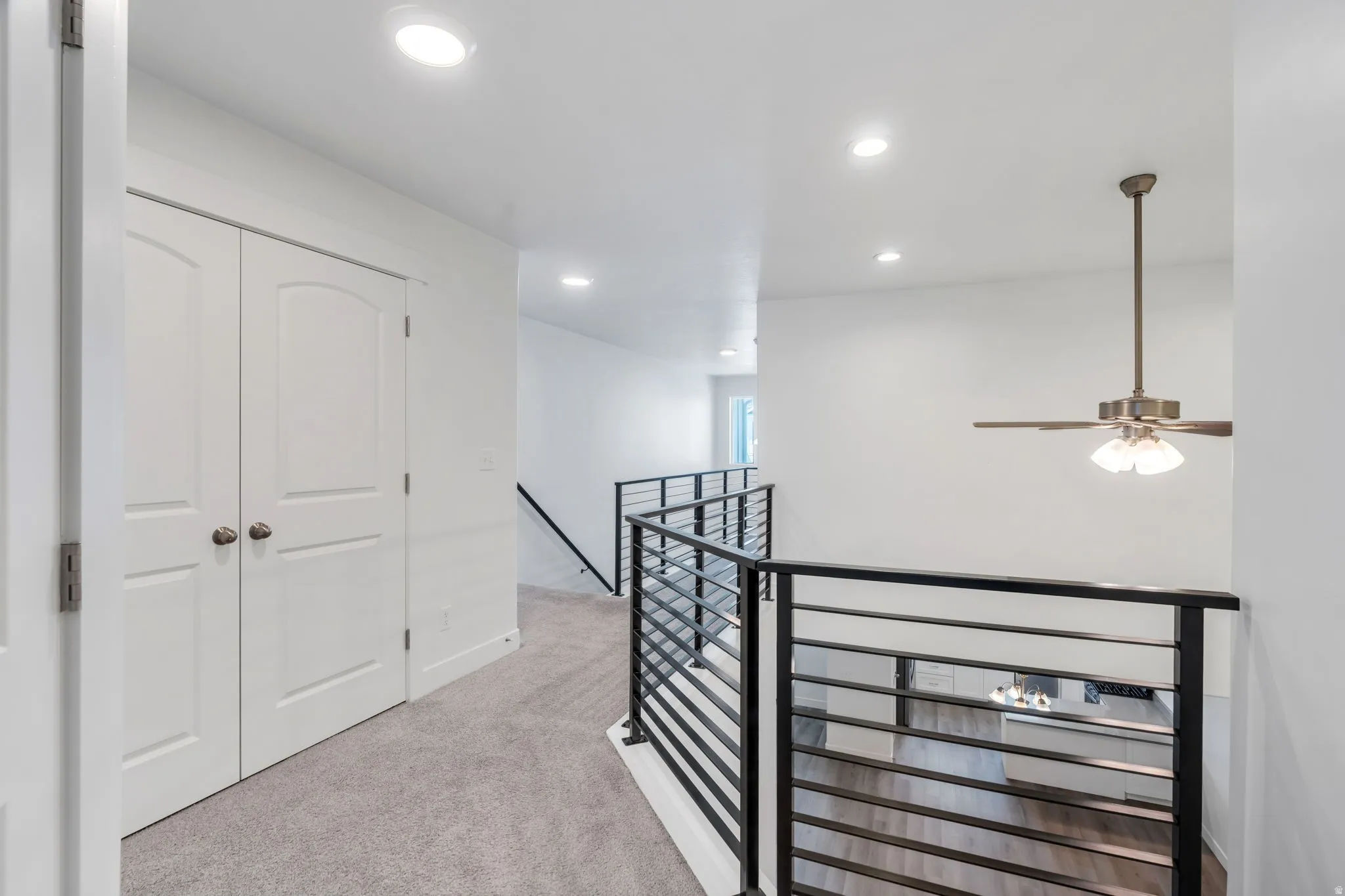 Upper-level landing featuring light gray carpeting, recessed ceiling lighting, a ceiling fan with integrated lighting, modern black horizontal bar railings, and white panel doors with arched tops