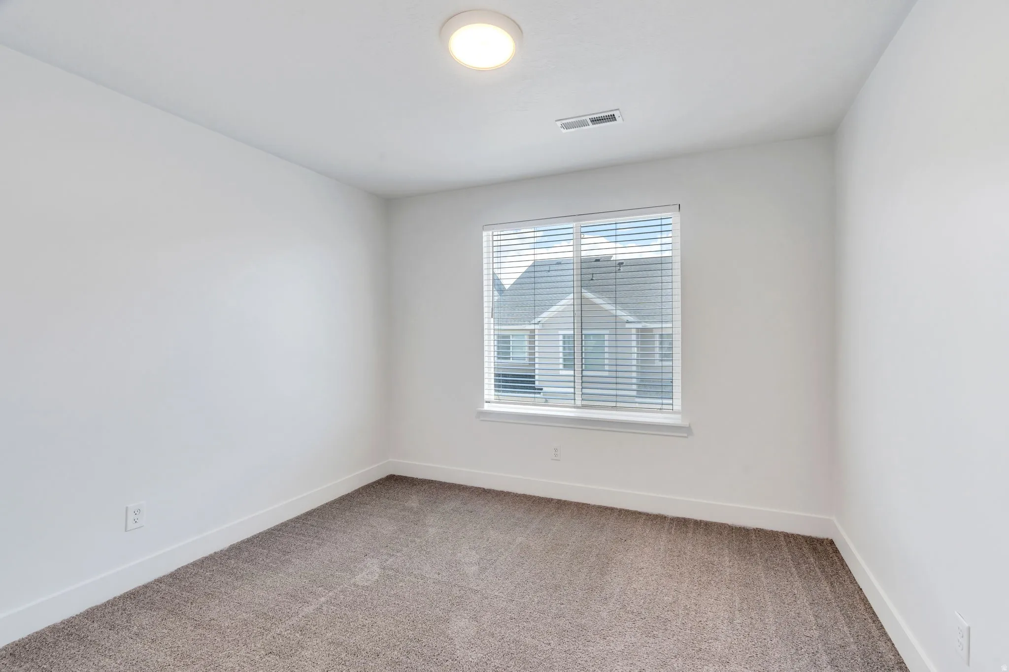 Bright room featuring a large window with blinds, neutral-toned carpet flooring, white walls, and a ceiling-mounted light fixture