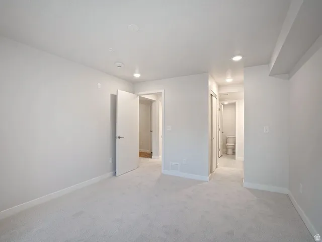 Spacious room featuring light gray carpeting, recessed lighting, white baseboards, a white interior door with brushed nickel hardware, and a separate hallway with tile flooring
