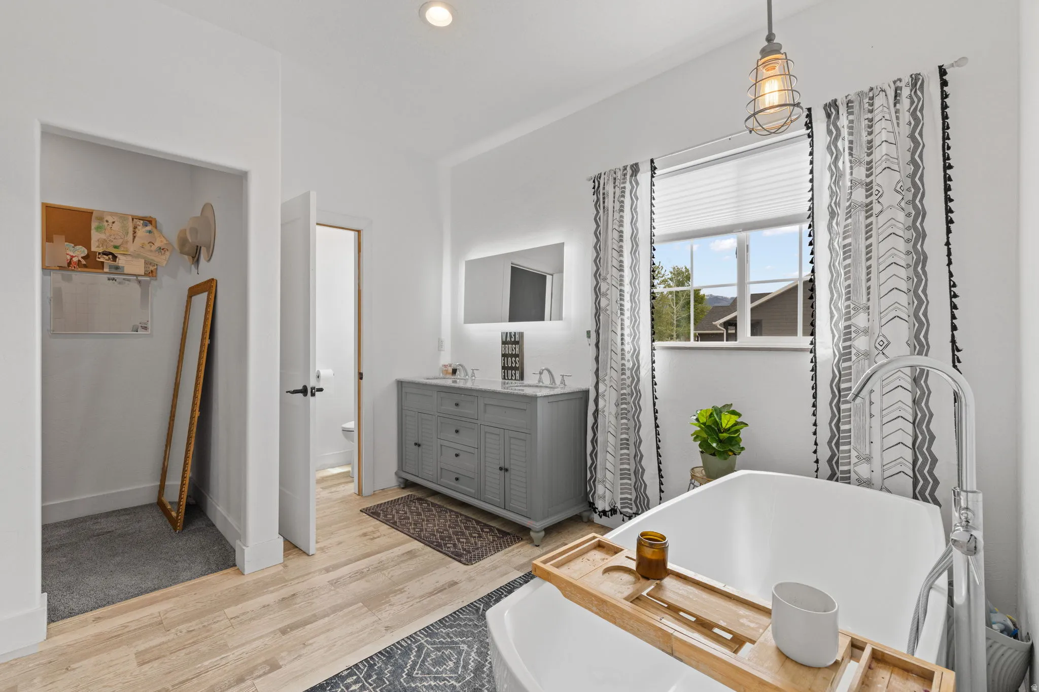 Primary Bedroom with en-suite, freestanding soaking tub with a floor-mounted faucet, wood-finish flooring, a double vanity with a light gray cabinet base, an illuminated mirror, and an open closet space