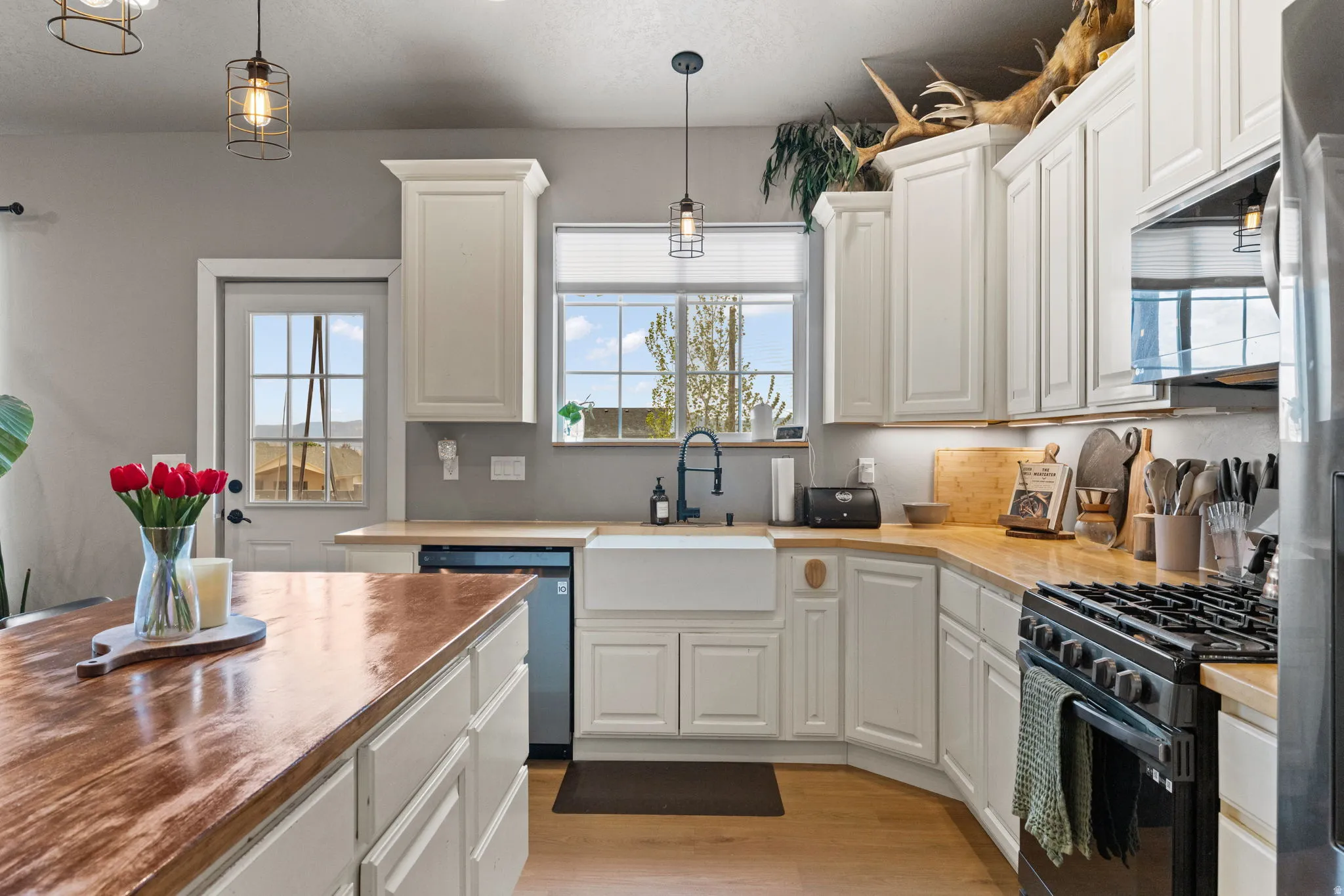 Kitchen featuring light wood-finish flooring, white cabinetry, wood-finish countertops, a farmhouse-style sink with a matte black faucet, and pendant lighting