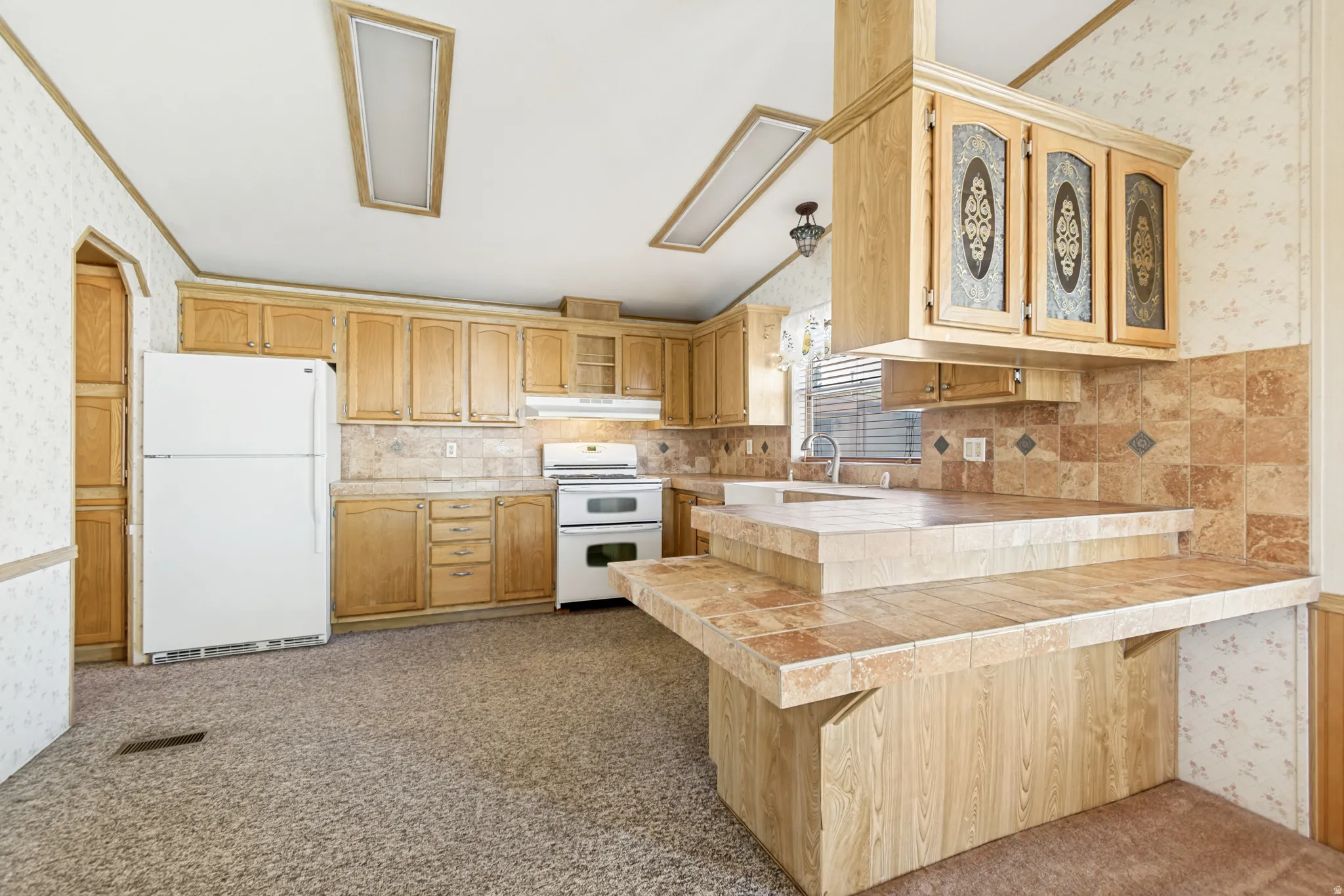 Kitchen featuring wallpapered walls, a peninsula, white appliances, a kitchen breakfast bar, and dark colored carpet