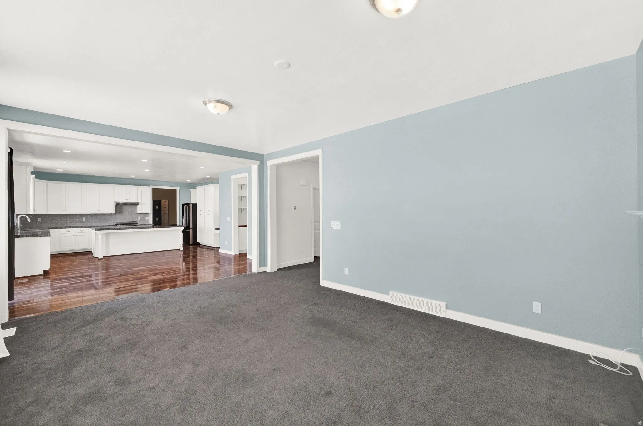 Spacious room with plush gray carpeting, leading to a kitchen featuring wood-finish flooring, white cabinetry, a central island, and recessed lighting