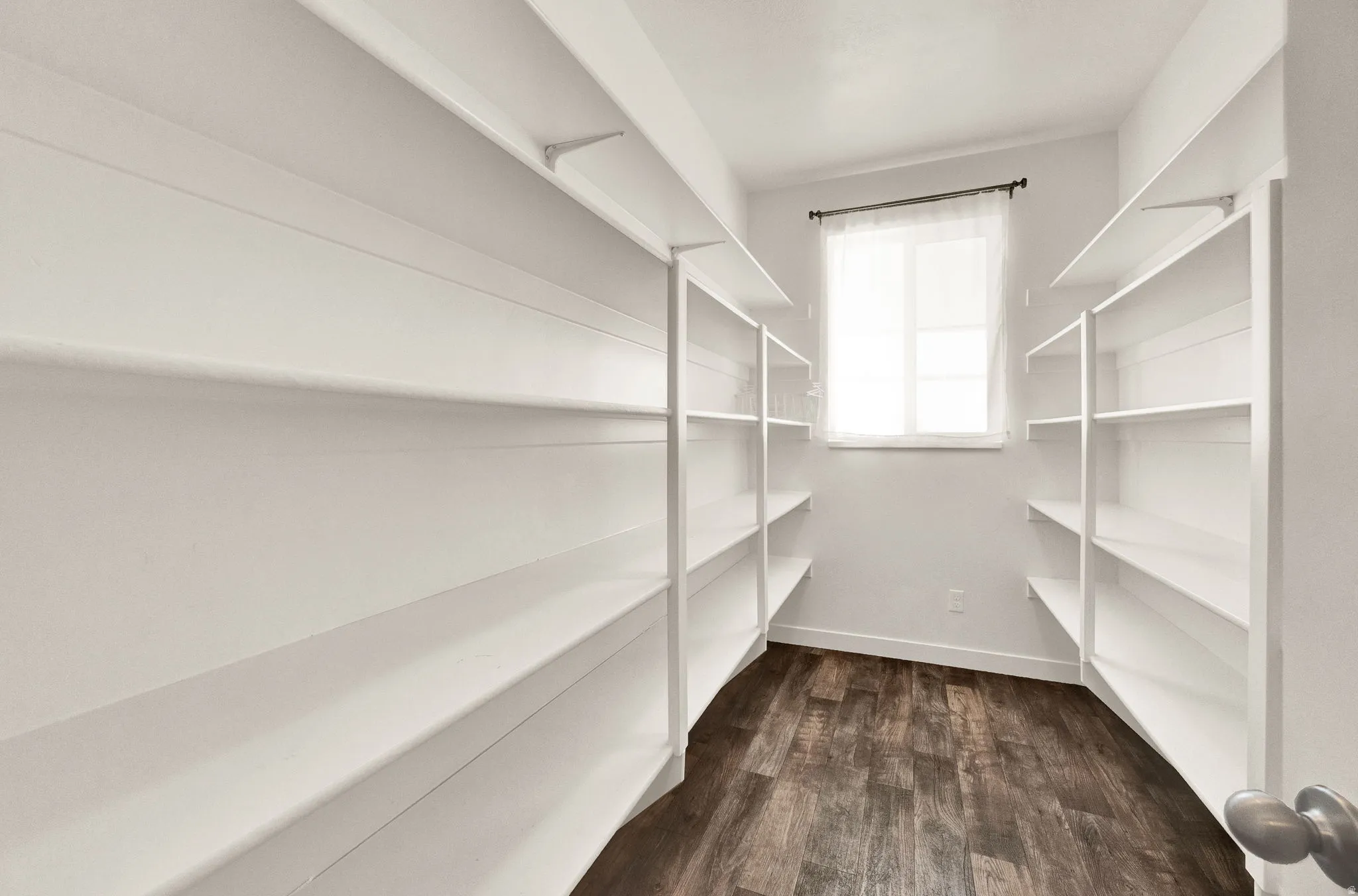 Spacious walk-in pantry featuring extensive white shelving, a window with natural light, and wood-finish flooring