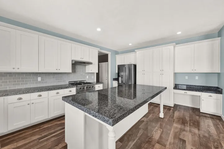Spacious kitchen featuring an expansive center island with a dark granite countertop, a stainless steel range, white shaker cabinetry, an integrated desk area, and wood-finish flooring